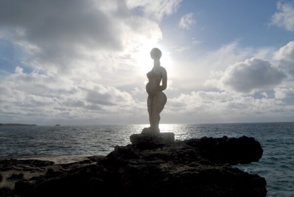 escultura de cuerto entero de una mujer con las manos bajo el vientre junto al Mediterrano, el sol se refleja en el agua